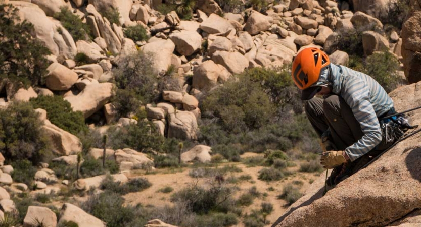 A person wearing safety gear crouches on a rock above a desert landscape covered with boulders. 